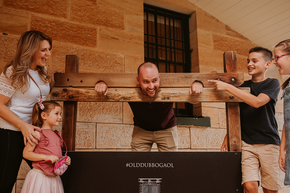 A man in the stocks at Old Dubbo Gaol, surrounded by his family laughing